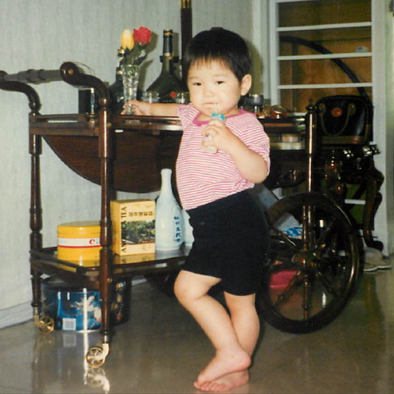 A chubby 2-year-old Eunice sipping yogurt and leaning against a bar cart with flowers in hand.